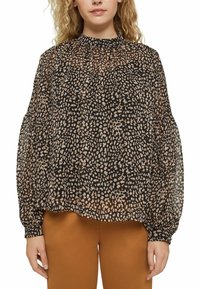 Blusa nera trasparente con scollatura arricciata, caratterizzata da un motivo a pois in tonalità beige e maniche lunghe voluminose, abbinata a pantaloni marroni.