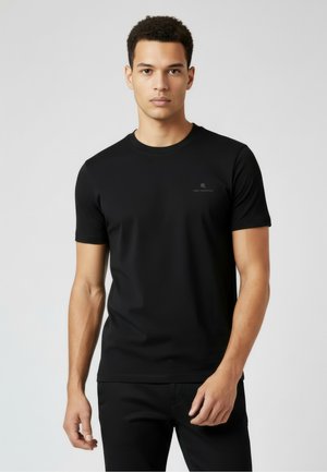 Nuori mies, jolla on lyhyet kiharat hiukset ja joka pukeutuu yksiväriseen mustaan lyhythihaiseen T-paitaan, seisoo harmaata taustaa vasten.