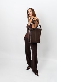 Brun lædertaske med et struktureret design, der har lodrette striber og to skulderremme, båret af en model i mørkt tøj.