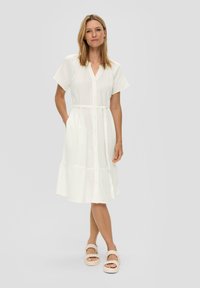 Witte kraagloze jurk met korte mouwen, gestructureerde stof, strikband in de taille en gelaagde zoom. Gecombineerd met beige gevlochten sandalen.
