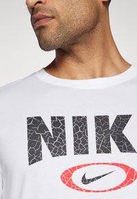 Hvit t-skjorte med korte ermer som har svarte sprukne bokstaver med teksten "NIK" og en rød oval med svart Nike-logo. Laget av bomull, klassisk rund halsdesign.