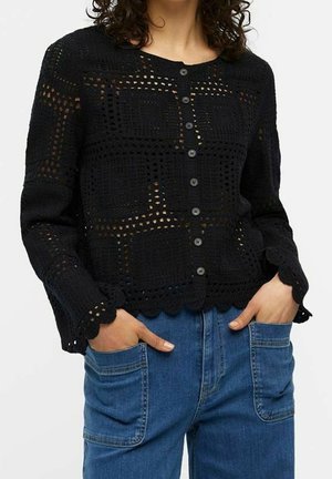 Frau trägt eine schwarze, gehäkelte Strickjacke mit langen Ärmeln und Knöpfen sowie blaue Jeans, die Hände in den vorderen Taschen.