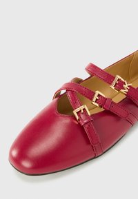 Rdeče usnjene čevlje s zaobljenim nosom, ki imajo križne trakove in zlate zaponke. Gladka tekstura z rahlim sijajem in izrezljanimi detajli.