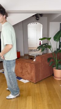 Hnědá kordurová pohovka, dřevěná podlaha, zelená rostlina v květináči, světle šedé stěny, vzorovaný koberec a postava v neformálním oblečení stojící v blízkosti.