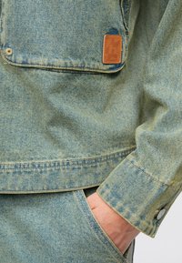 Φωτεινό μπλε τζιν μπουφάν με τσέπη στο στήθος, καφέ δερμάτινη ετικέτα και μεταλλικά κουμπιά. Υφή υφάσματος με ξεθωριασμένα σχέδια.