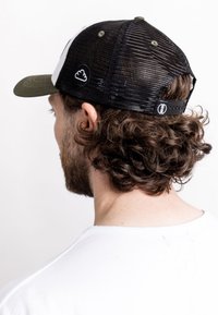 Black mesh trucker cap with a green bill, featuring a white logo patch and adjustable snap closure at the back. Curly hair visible.