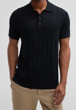 Homme portant un polo en maille noir à manches courtes avec des rayures texturées verticales et un pantalon beige, debout avec une main dans la poche.