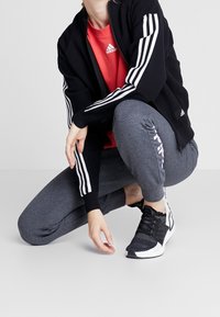 Chaqueta deportiva negra con rayas blancas, camiseta roja, pantalones jogger grises con acento de logo y zapatillas negras/blancas. Diseño elegante y deportivo.