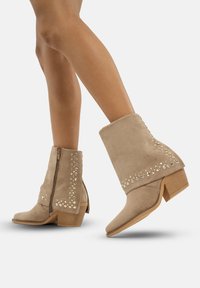 Botas de tobillo de ante beige con tacón apilado, que presentan un patrón decorativo de tachuelas a lo largo del costado y un cierre de cremallera lateral.