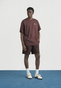 Νεαρός άνδρας στέκεται φορώντας σκούρο καφέ μπλουζάκι Adidas, ταιριαστά σορτσάκια, λευκές κάλτσες και λευκά αθλητικά παπούτσια Adidas σε μπλε δάπεδο.