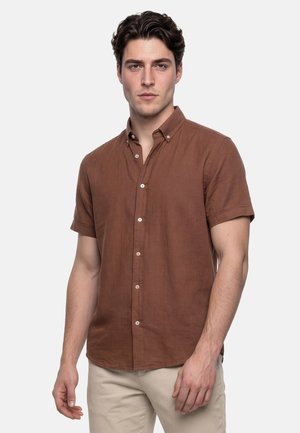 Joven con cabello oscuro y ondulado, que lleva una camisa marrón de manga corta abotonada y pantalones beige, de pie frente a un fondo liso.
