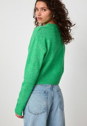 Junge Frau mit lockigem Haar, die einen leuchtend grünen Pullover und hellblaue hochtaillierte Jeans trägt und über ihre Schulter gegen einen schlichten Hintergrund schaut.