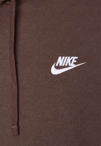 Brauner Hoodie aus weichem Material mit einem auffälligen weißen, gestickten Nike-Logo auf der Brust und einem Kordelzug an der Kapuze.