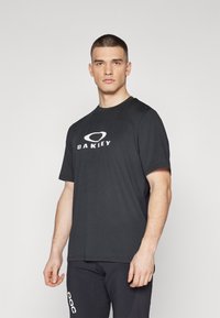 Crna majica kratkih rukava s okruglim izrezom, koja na prednjoj strani ima bijeli "Oakley" logo i teksturiranu površinu.