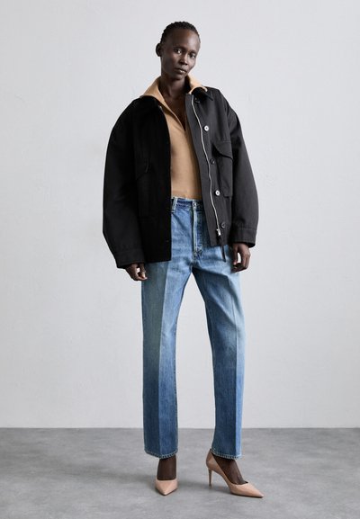 Black oversized jacket with front pockets, beige shirt underneath, light blue straight-leg jeans, and nude high-heeled shoes.