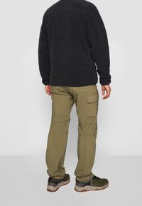 Giacca in pile nera, con una superficie testurizzata. Pantaloni cargo verdi oliva con tasche laterali. Calzature di colore verde scuro con accenti grigi.