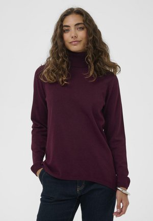 Junge Frau mit lockigem braunem Haar, die einen dunkelvioletten Rollkragenpullover und dunkle Jeans trägt und mit einer Hand in der Tasche steht.