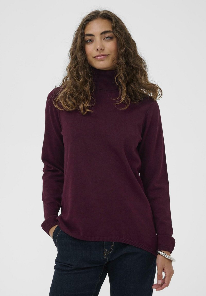 Mlada ženska z kodrastimi rjavimi lasmi, oblečena v temno vijolični pulover s turtleneck izrezom in temne kavbojke, stoji z eno roko v žepu.
