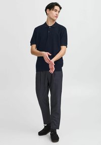 Kék póló ing gallérral és két gombbal, szürke pliszírozott nadrággal párosítva. A modell összekapcsolt kezekkel áll, fekete cipőt viselve.