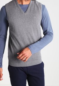 Pull sans manches en laine grise tricotée avec un col en V, porté sur une chemise à manches longues bleu clair. Texture lisse, coupe décontractée.