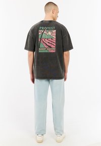 Grijze oversized t-shirt met een kleurrijk graphic design op de achterkant met de teksten "PROTEST" en "SUNSET SESSIONS," gecombineerd met lichtblauwe jeans.