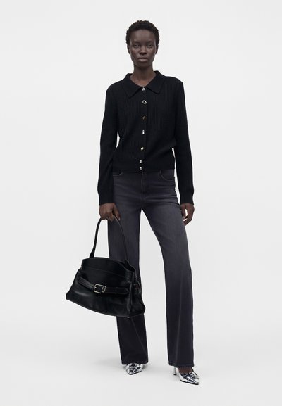 Femme portant un cardigan noir boutonné et un jean foncé, tenant un grand sac à main noir, debout devant un fond blanc.