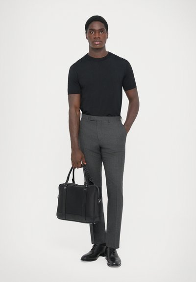 Camiseta negra de manga corta, pantalones grises a medida y zapatos de cuero negros. Sosteniendo un bolso de cuero negro, de pie contra un fondo blanco.