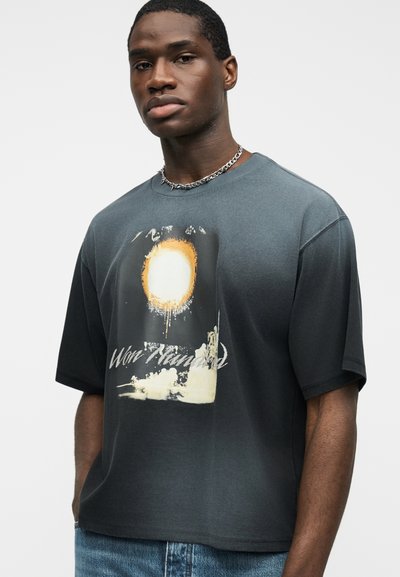 Νεαρός άνδρας που φοράει σκούρο γκρι μπλουζάκι με κίτρινο ήλιο και τη φράση "Won't Handed", συνδυασμένο με μπλε τζιν και ασημένιο αλυσίδα κολιέ.