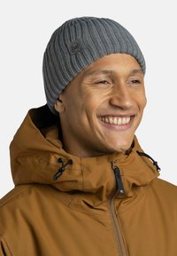 Gorro de punto gris con textura acanalada y parche de logo, llevado por una persona con una chaqueta marrón con capucha ajustable y cremallera.