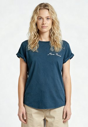 Blonde vrouw met golvend haar, die een marineblauw t-shirt draagt met opgerolde mouwen en de tekst "Mira Paris" en beige broek, kijkt recht vooruit met een neutrale gezichtsuitdrukking.