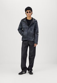 Zwarte, waterdichte jas met een gestructureerd patroon, ritsluiting en verstelbare capuchon. Gecombineerd met donkere jeans en zwarte sneakers.