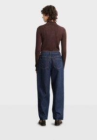 Dunkelbrauner gerippter Pullover mit hohem Kragen, kombiniert mit lässigen, dunkelblauen Jeans. Auffällige Rückentaschendetails. Schwarze Schuhe.