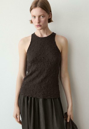 Jeune femme aux cheveux courts portant un haut sans manches texturé de couleur sombre et tenant un tissu foncé contre un mur clair uni.