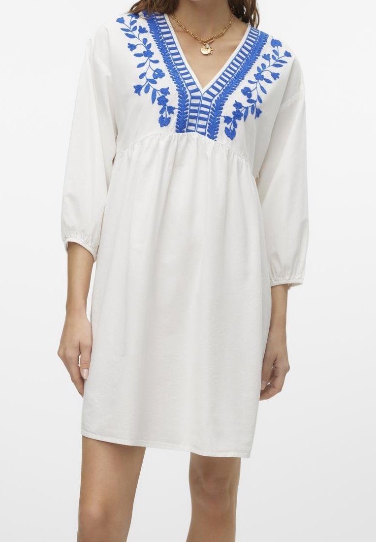 Vit bomullsklänning med blå blommönster och randiga detaljer längs V-halsen. Har trekvartslånga ärmar och en avslappnad passform.
