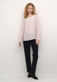 Lys pink langærmet bluse med delikat blondedetalje, afslappet pasform og volangmanchetter, parret med mørke denimbukser og sorte hælede støvler.
