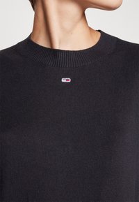 Gros plan sur une personne portant un pull ras du cou noir côtelé avec un petit logo rectangulaire rouge, blanc et bleu marine centré sous l'encolure.