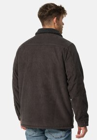 Donkergrijze corduroy jas met een gestructureerde shearling kraag, voorzien van een knoopsluiting en zichtbare stikdetails op de achterkant.