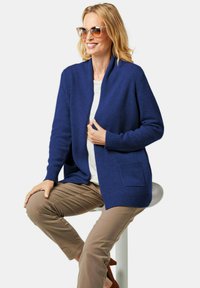 Cardigan tricoté bleu avec un col châle, des poignets côtelés et une poche avant. Porté par-dessus une chemise blanche, associé à un pantalon marron clair.