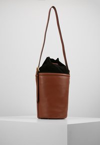 Borsa a secchiello in pelle marrone con chiusura a cordoncino in tessuto nero e lunga tracolla, esposta su un piedistallo bianco.