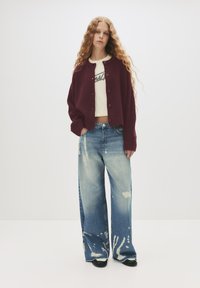 Cardigan lavorato in borgogna con bottoni, t-shirt corta bianca con grafica e jeans blu a effetto acido con macchie di candeggina, a vestibilità ampia.