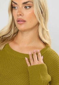 Oliivinvihreä neulepaita, jossa on leveä kaula-aukko, ribattuja hihansuita ja teksturoitu kangas. Käsi on sijoitettu hellästi olkapäälle, korostaen materiaalia.