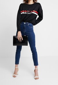 Sweat-shirt noir avec des rayures rouges et blanches, jean skinny bleu, et pochette noire à texture crocodile. Sandales blanches à brides.