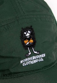 Vihreä lakki, jonka etuosassa on musta, karvainen sarjakuvahahmo kirjailtuna, pitäen oransseja esineitä, ja alla valkoinen teksti "NORDICBUDDIES CLOTHING CO."