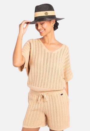 Kvinne som smiler, iført beige kortermet strikket topp og shorts, justerer en svart og beige hatt med bred brem og dekorativ pin.