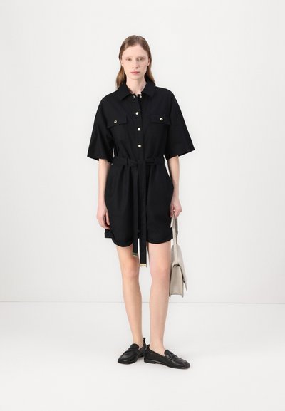 Robe noire à manches courtes avec un devant à boutons, deux poches poitrine et une ceinture en tissu. Portée avec des chaussures noires et un sac à main de couleur claire.