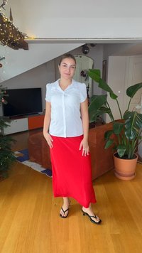 Λευκό πουκάμισο με κοντά μανίκια και κουμπιά, με γιακά, σε συνδυασμό με μακρύ κόκκινο φούστα. Φορεμένο με μαύρα σαγιονάρες σε ξύλινο πάτωμα. Κοντά υπάρχει πράσινο φυτό.