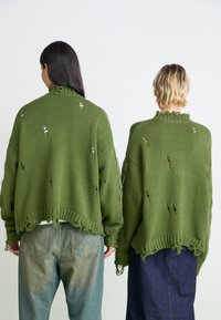 Pulls en maille verte surdimensionnés avec des bords effilochés et des détails usés, dotés d'un col haut et de manches longues, associés à un jean bleu.