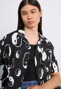 Chemise noire à manches courtes avec un motif yin-yang blanc, des boutons sur le devant et une poche poitrine. Tissu lisse avec une coupe décontractée.