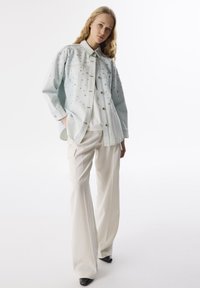 Camicia di jeans azzurro chiaro con dettagli decorativi, abbinata a pantaloni a gamba larga color crema. La camicia presenta una chiusura con bottoni e un colletto.