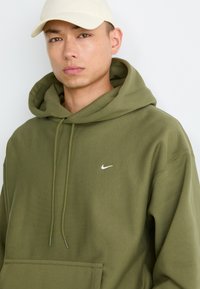 Ung mand iført en beige kasket og en olivengrøn Nike hoodie med snore, som står foran en ensfarvet lys baggrund.